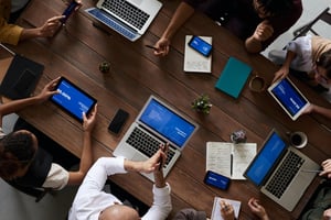 Top‑down view of a team collaborating at a table with laptops, tablets, notes, and shared data screens.