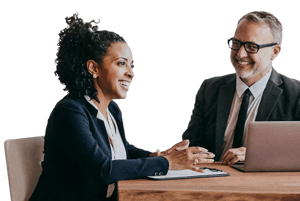 Two business professionals seated at a desk, discussing documents and working on a laptop during a meeting.