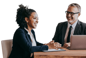 Two business professionals seated at a desk, discussing documents and working on a laptop during a meeting.